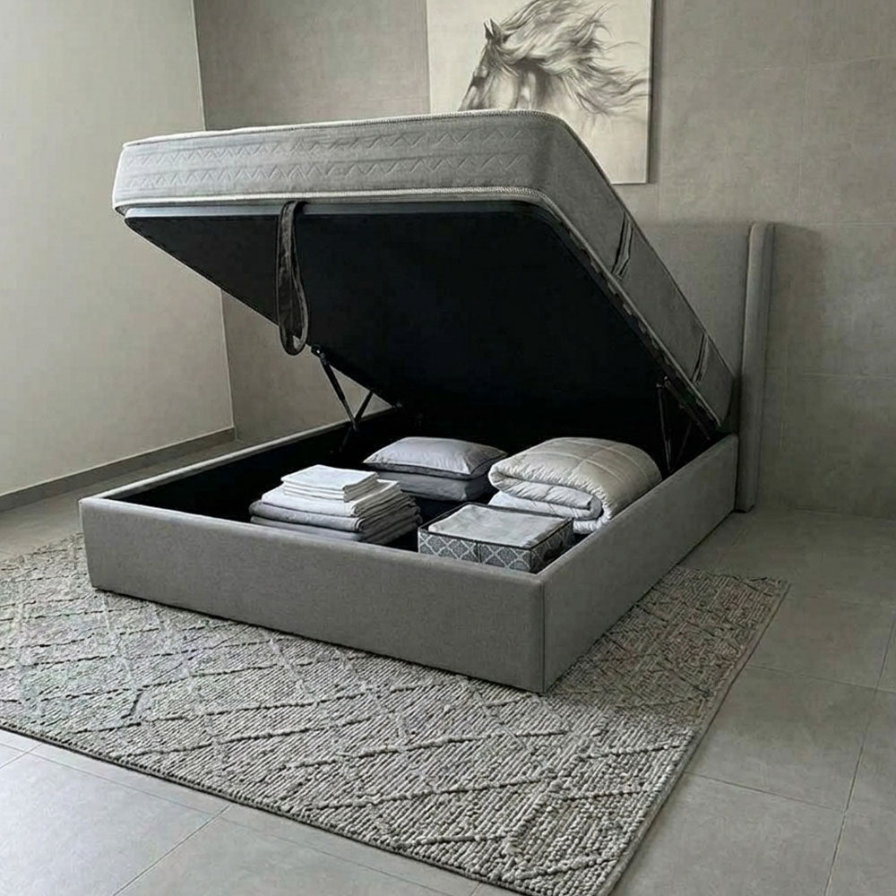 Gray bed with storage compartment open, revealing folded bedding and pillows on a neutral-colored floor.