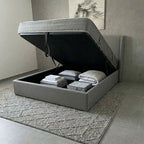 Gray bed with storage compartment open, revealing folded bedding and pillows on a neutral-colored floor.