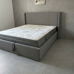 Gray upholstered bed with a mattress in a room with tiled flooring and a neutral wall.