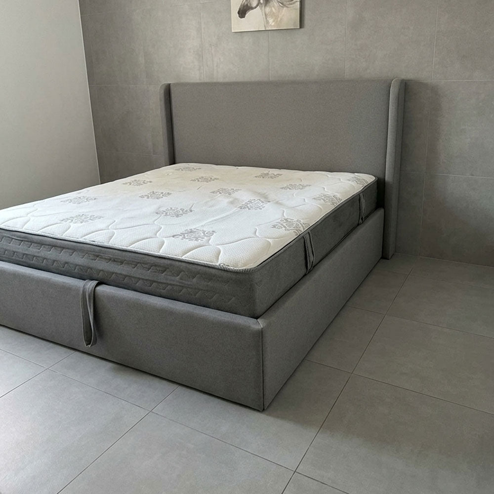 Gray upholstered bed with a mattress in a room with tiled flooring and a neutral wall.