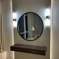 Round mirror with a dark frame on a wall with two lights, below a window with curtains.