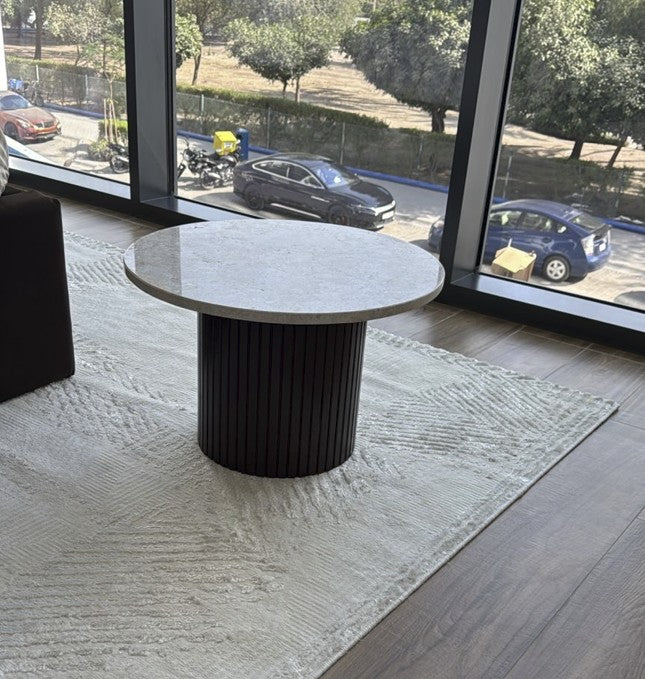 Round table with a marble top and black cylindrical base in a room with large windows showing cars outside.